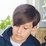 Nærbilde av en gutt med mørkt hår og blå hettegenser som står inne på et kjøkken og ser nedover.