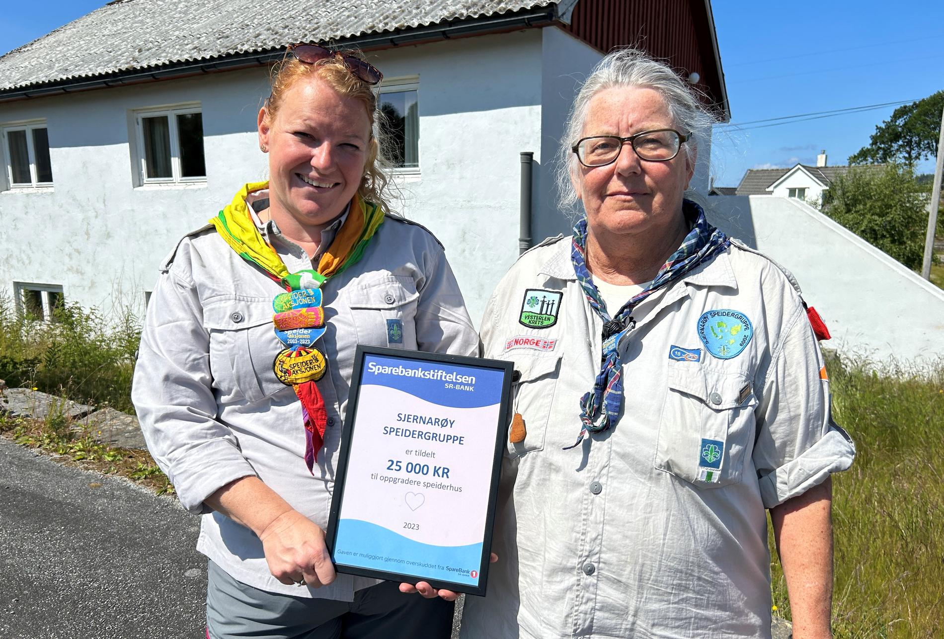 Gruppeleiarar Tove Bergjord Tysdal (t.v.) og Rita Vik i Sjernarøy Speidergruppe føre klubbhuset sitt på Nord-Talgje og med pengegåva frå Sparebankstiftelsen SR-Bank.
