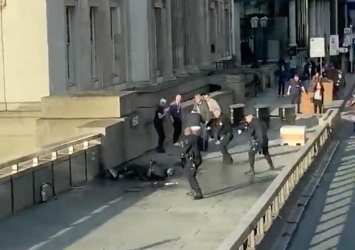 Bildet er en grabb fra en video, som viser terroristen omgitt av tre politimenn. Bak står de fire mennene som overmannet ham. 